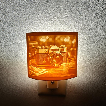 Warm glowing camera lithophane night light featuring a detailed vintage-style camera on a tabletop with blurred background lights, curved photography-themed decor for bedrooms, offices, studios, or camera lovers.