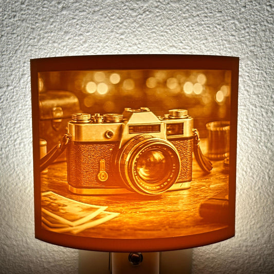 Warm glowing camera lithophane night light featuring a detailed vintage-style camera on a tabletop with blurred background lights, curved photography-themed decor for bedrooms, offices, studios, or camera lovers.